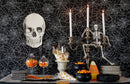 A Halloween-themed tabletop setup with a black spiderweb-patterned backdrop, a large white skull decoration, a skeleton figurine on a candelabra, white taper candles, black bowls, plates, striped napkins, goblets filled with orange and yellow candy corn, and a striped table runner.