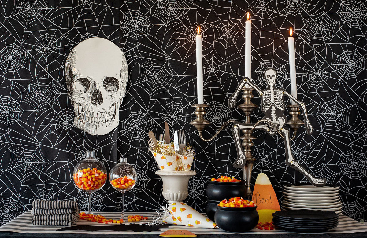 A Halloween-themed tabletop setup with a black spiderweb-patterned backdrop, a large white skull decoration, a skeleton figurine on a candelabra, white taper candles, black bowls, plates, striped napkins, goblets filled with orange and yellow candy corn, and a striped table runner.