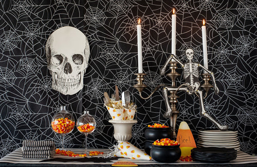 A Halloween-themed tabletop setup with a black spiderweb-patterned backdrop, a large white skull decoration, a skeleton figurine on a candelabra, white taper candles, black bowls, plates, striped napkins, goblets filled with orange and yellow candy corn, and a striped table runner.