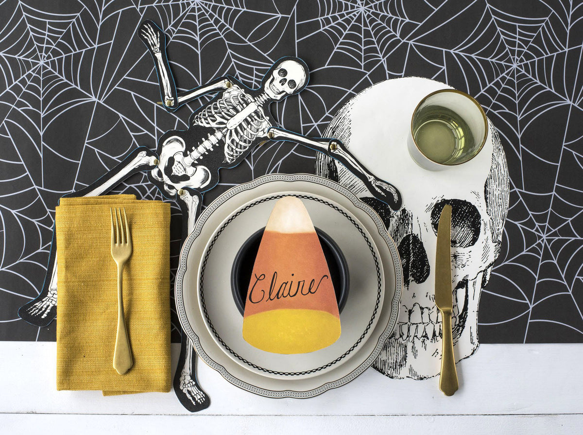 A styled Halloween table setting featuring a central place setting with a candy corn plate, a yellow napkin with a gold fork, a skull-shaped placemat, skeleton cutouts, and a spiderweb-patterned backdrop.
