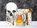 A styled Halloween table setting featuring a large black-and-white skull print on a white placemat, a white napkin folded with a candy corn centerpiece, gold-toned flatware, a glass of amber liquid, and scattered candy corn motifs on a black spiderweb-patterned background.
