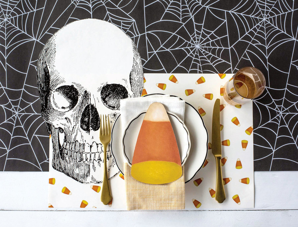 A styled Halloween table setting featuring a large black-and-white skull print on a white placemat, a white napkin folded with a candy corn centerpiece, gold-toned flatware, a glass of amber liquid, and scattered candy corn motifs on a black spiderweb-patterned background.