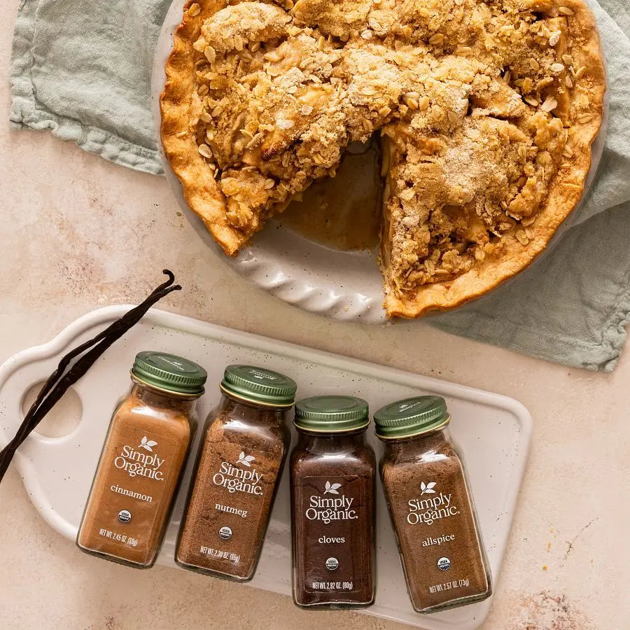 Apple pie with a slice missing, with four bottles of Simply Organic spices placed on a white plate.