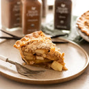 Slice of apple pie on a plate with a fork, against a blurred background of coffee jars.