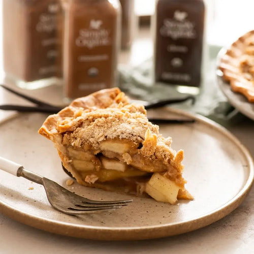Slice of apple pie on a plate with a fork, against a blurred background of coffee jars.