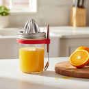 Glass jar filled with orange juice with a metal citrus press on top and red silicone spoon holder with a spoon attached on a kitchen counter with oranges nearby.
