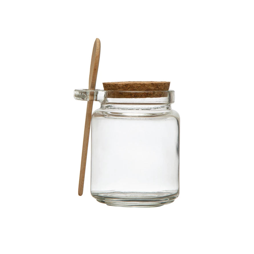 A clear glass jar with a cork lid and a wooden spoon attached to the side.