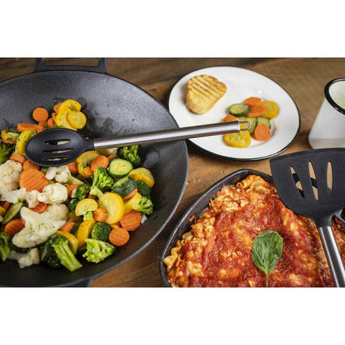 A slotted spoon with a silver handle in a pan full of mixed vegetables, next to another pan full of food with a slotted spatula and a plate and cup nearby on a wooden surface.