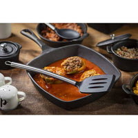 A slotted spatula with a stainless steel handle sitting on a cast iron pan with food in it atop a wooden table with other pots, pans, and pepper shaker.