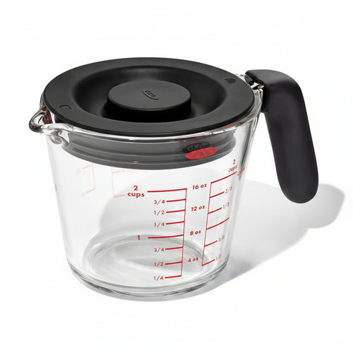 A clear glass measuring cup with black lid and a red measurement scale on the side, with a black handle and a brand logo on the front.