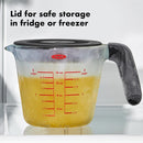 A glass measuring cup with a lid holding yellow liquid in a freezer with frost on it and text noting that the lid is safe for the fridge or freezer.