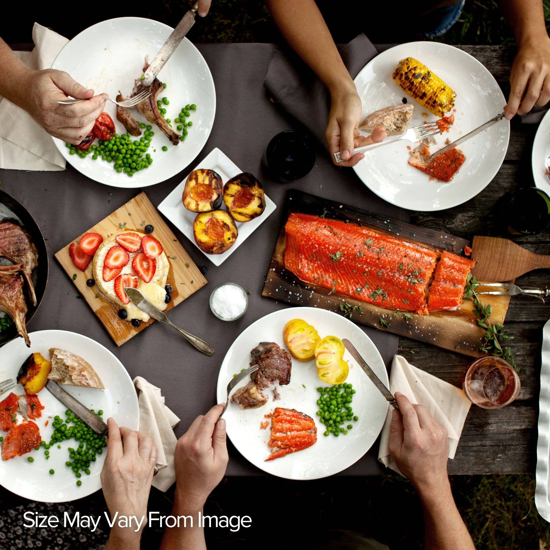 People enjoying a meal around a table with various dishes including salmon resting on a grilling plank and various vegetables.