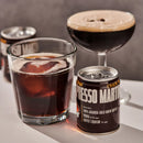 A can of Espresso Martini coffee liqueur in the foreground, with a glass of dark iced coffee and a coupe glass of dark coffee with foam behind it on a tabletop against a gray background.