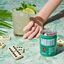 A hand holds a mojito glass with mint and lime beside a can of Mojito on a turquoise stone surface, with dominoes and palm leaves around it.