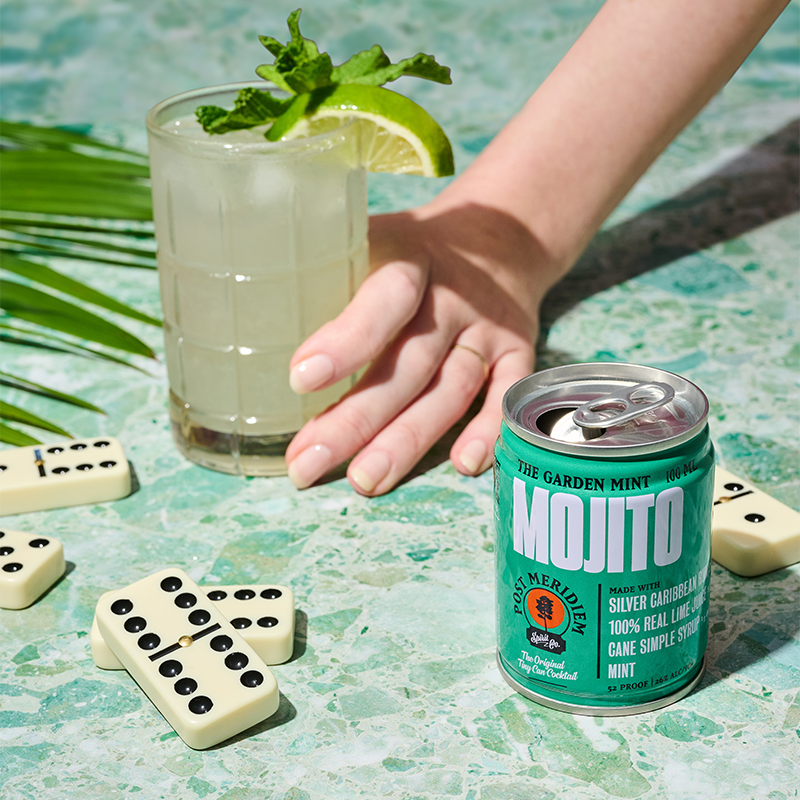A hand holds a mojito glass with mint and lime beside a can of Mojito on a turquoise stone surface, with dominoes and palm leaves around it.