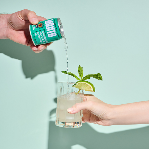 A hand pours a mojito from a green can into a glass with ice, mint, and a lime wedge, held by another hand against a solid mint green background.
