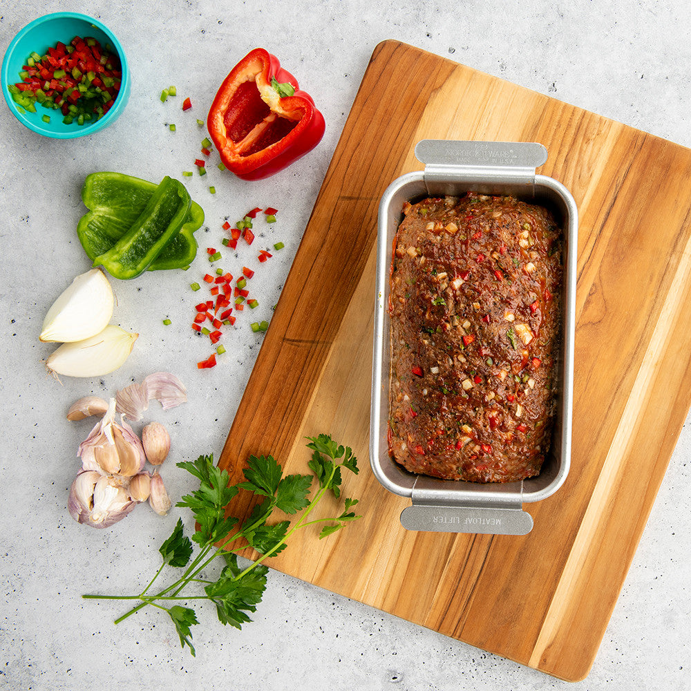 Loaf of meatloaf in a metal loaf pan on a wooden cutting board with vegetables and herbs.
