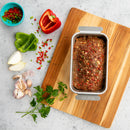 Loaf of meatloaf in a metal loaf pan on a wooden cutting board with vegetables and herbs.
