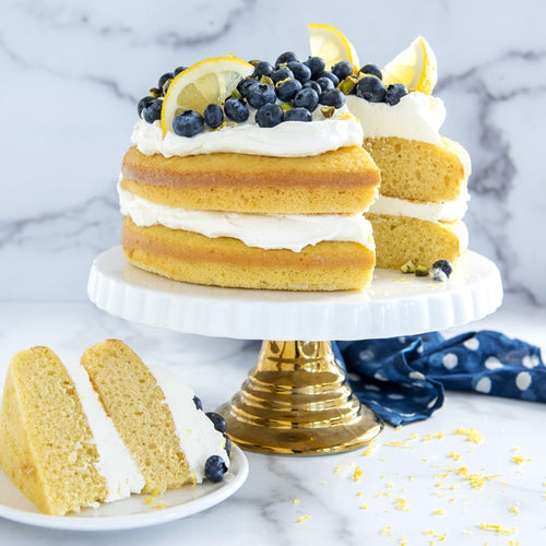 Two-layer lemon cake with blueberries on a marble surface, one slice cut and placed on a plate.
