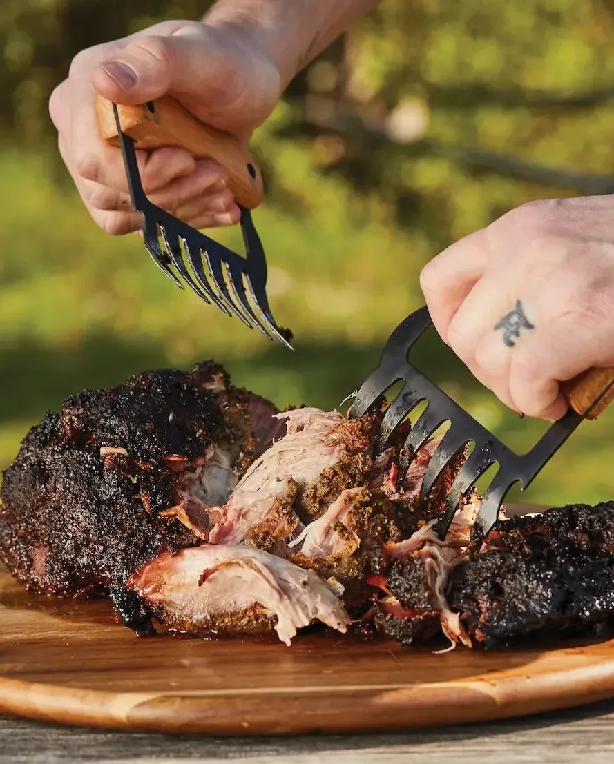 Person using meat shredding claws outdoors to shred barbecue on a wooden surface.