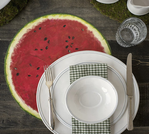 A rustic place setting featuring a bowl, plates, flatware, and a drinking glass with a watermelon paper placemat on a dark wooden surface.