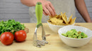 Hand picking up chips near a bowl of guacamole with a green and silver hand masher on a wooden surface with tomatoes and cilantro.
