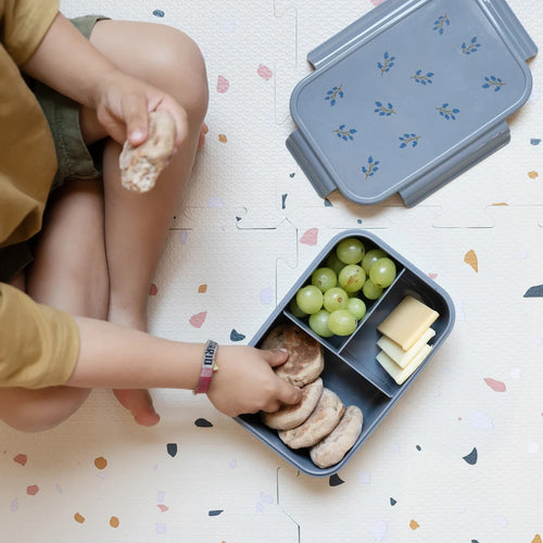 A child taking food out of a bento box while sitting on a patterned floor.