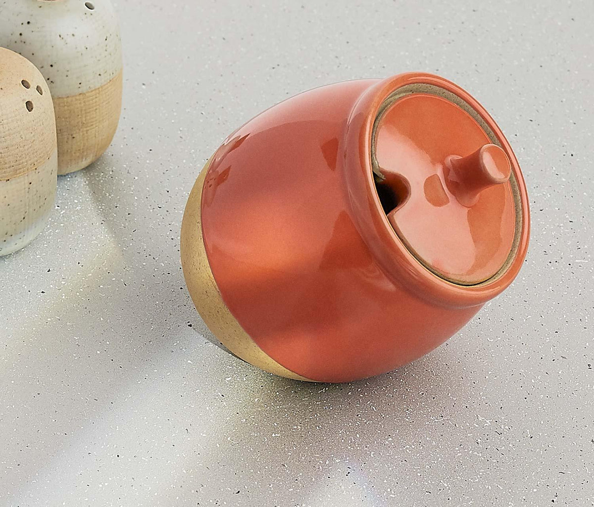 A glossy ceramic lidded container in a warm terracotta orange color with a natural beige base, photographed on a light speckled surface next to other ceramic items.