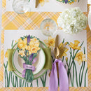Easter table setting with floral placemats, yellow flowers, and gold cutlery on a yellow checkered tablecloth.