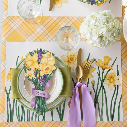 Easter table setting with floral placemats, yellow flowers, and gold cutlery on a yellow checkered tablecloth.