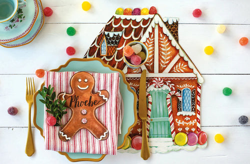 Decorative gingerbread house placemat and gingerbread man cookie table accent on a plate with colorful candies on a white wooden surface.