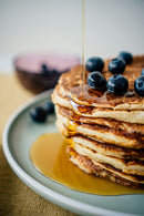 A close-up shot of a tall stack of golden-brown pancakes topped with fresh blueberries and a stream of syrup being poured over them, served on a light gray plate.