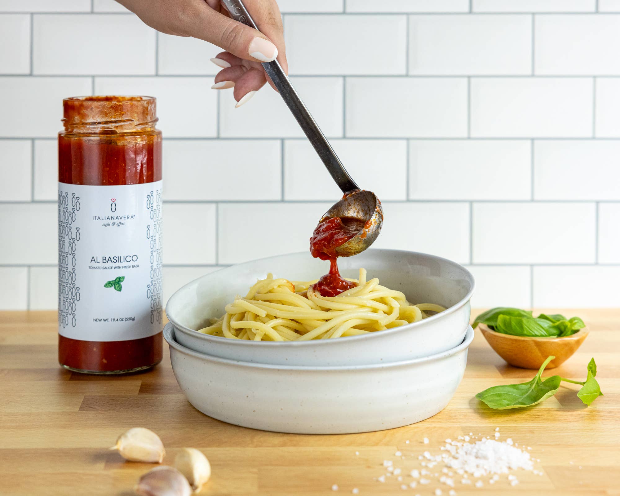 A person adding a ladle of sauce to a bowl of pasta next to a few leaves and cloves of garlic on a wooden surface.