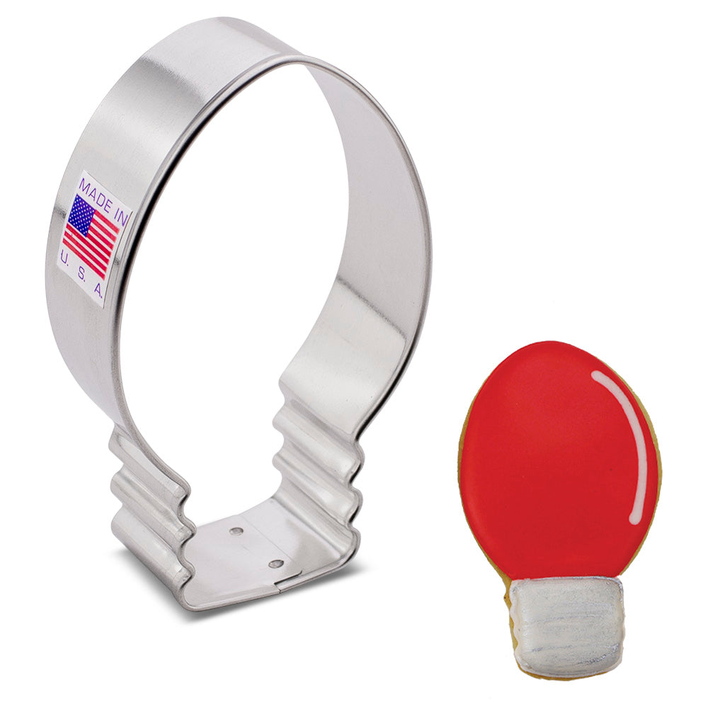 A cookie cutter made to look like a Christmas light bulb with an example cookie on a white surface.