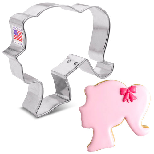 A metal cookie cutter in the shape of a doll's head, with a pink bow detail on the resulting cookie shape.