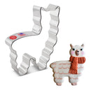 A stainless steel cookie cutter in the shape of a llama with a sticker stating it is made in the USA, next to a cookie decorated to look like a llama with a scarf.