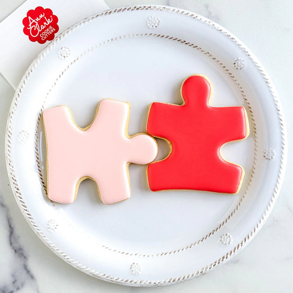 Two puzzle piece-shaped cookies, one pink and one red, on a white plate with 'Ann Clark' branding.