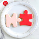 Two puzzle piece-shaped cookies, one pink and one red, on a white plate with 'Ann Clark' branding.