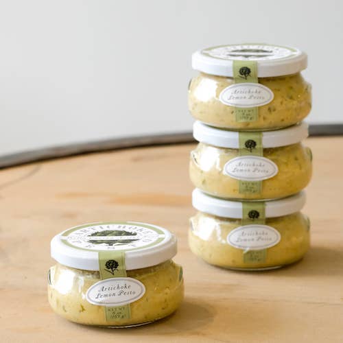 Four jars of artichoke lemon pesto on a wooden surface with a white background
