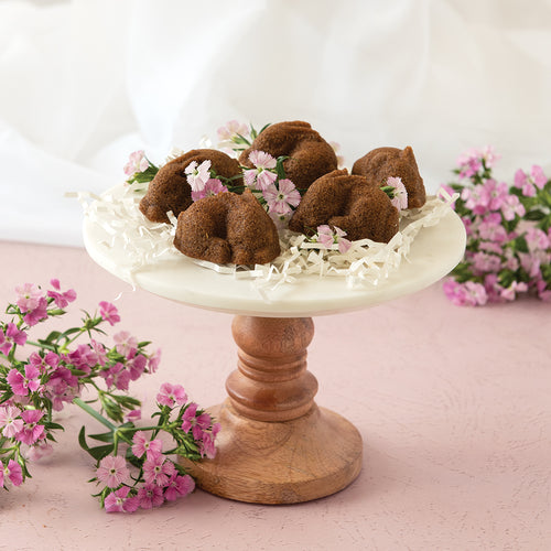 Decorative display of small brown bunny cakes on a white pedestal with pink flowers on a light pink background