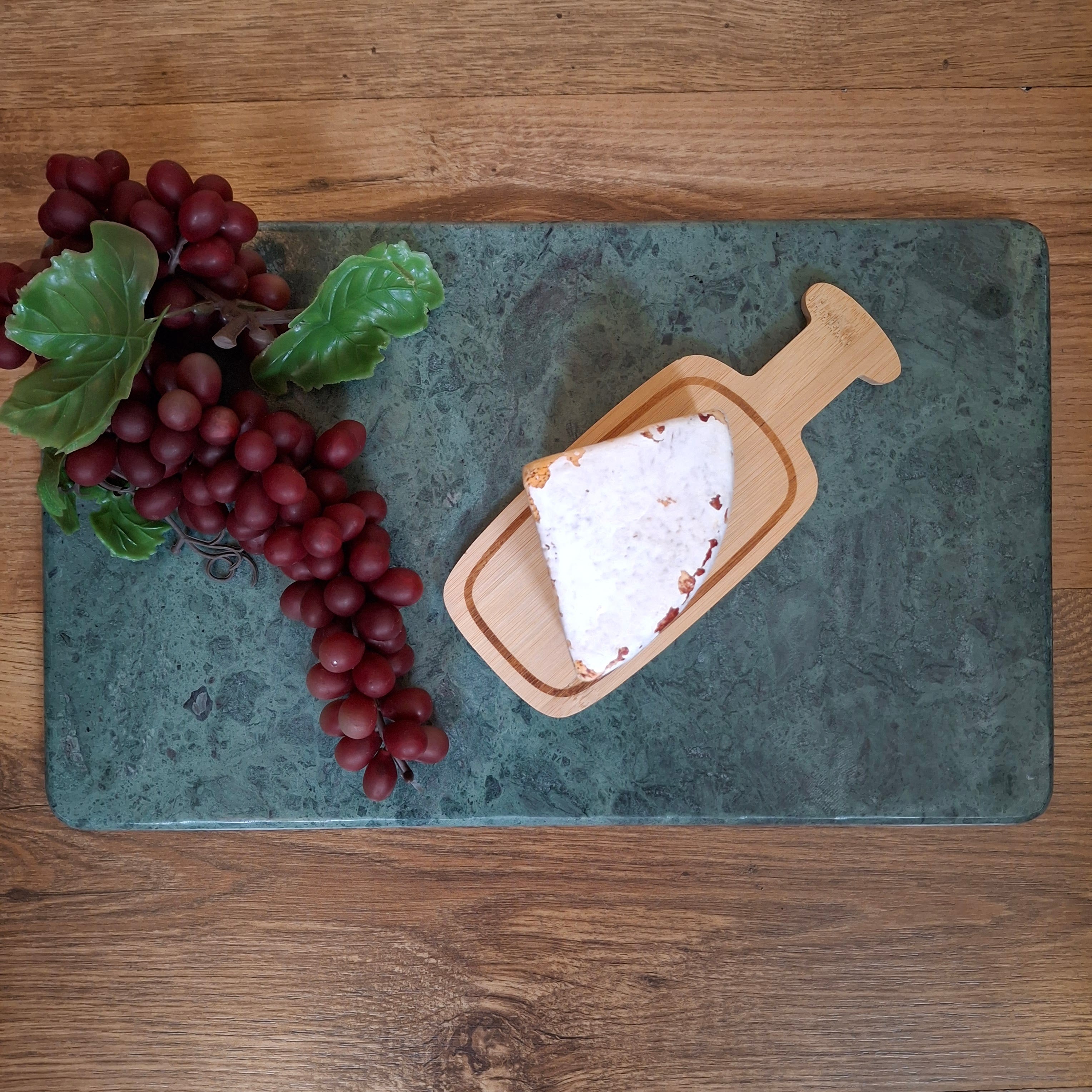 Sliced white cheese on a wooden board with red grapes on a green marble surface, all on a wooden table.