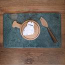 Chevre cheese on a wooden board with a cheese knife on a green marble surface, all on a wooden table.
