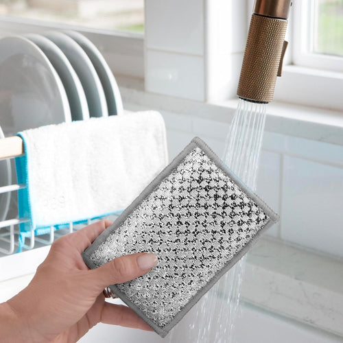 A hand holds a rectangular stainless steel cleaning sponge under a running faucet in a bright kitchen setting.
