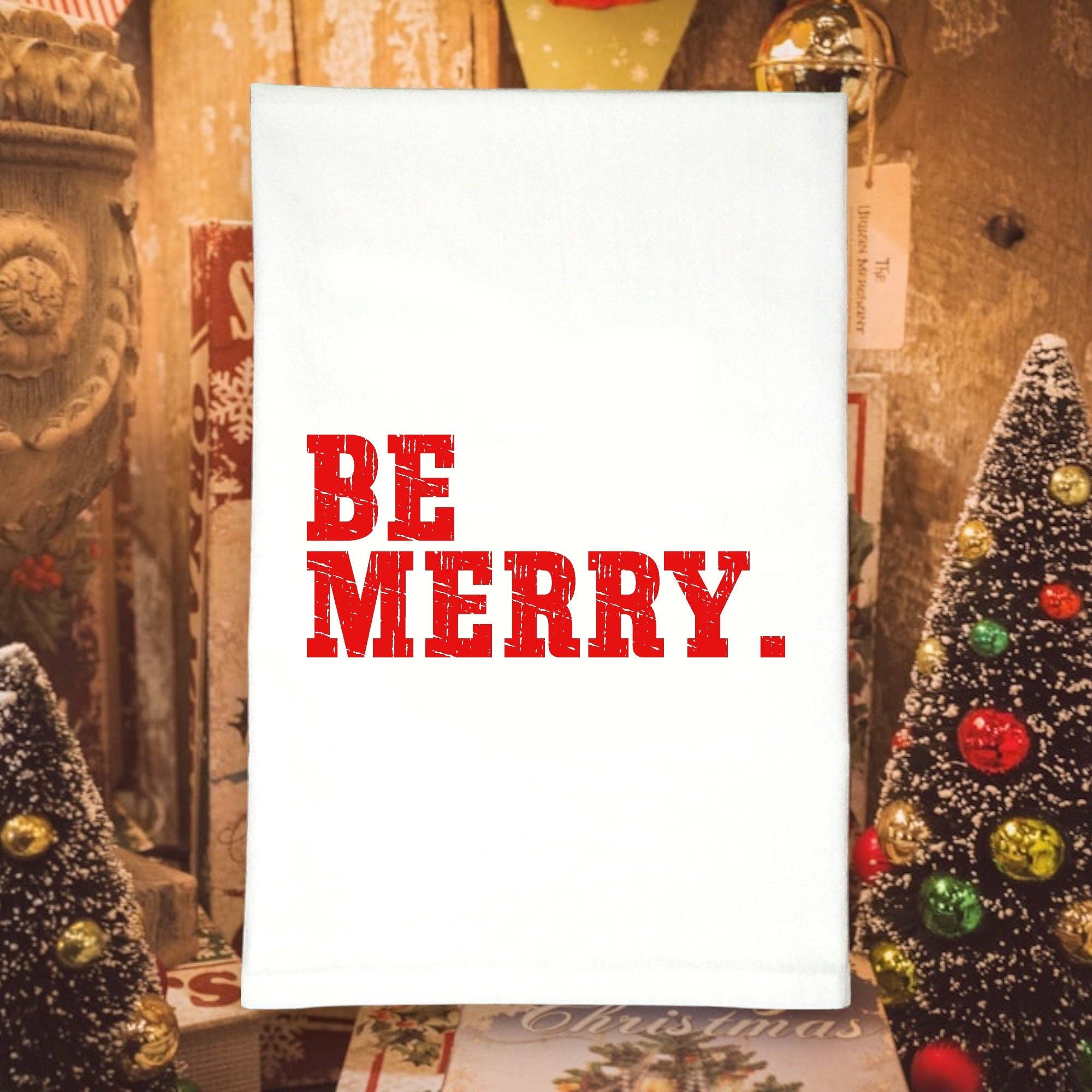 Kitchen towel with 'BE MERRY' text in red on a white background, surrounded by Christmas decorations.