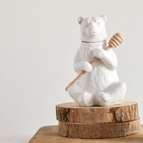 White ceramic bear figurine holding a bamboo honey dipper on a wooden base against a light gray background