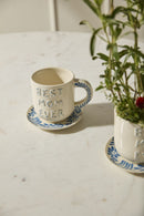 Ceramic mug with 'Best Mom Ever' text on a saucer, next to a plant on a light surface.