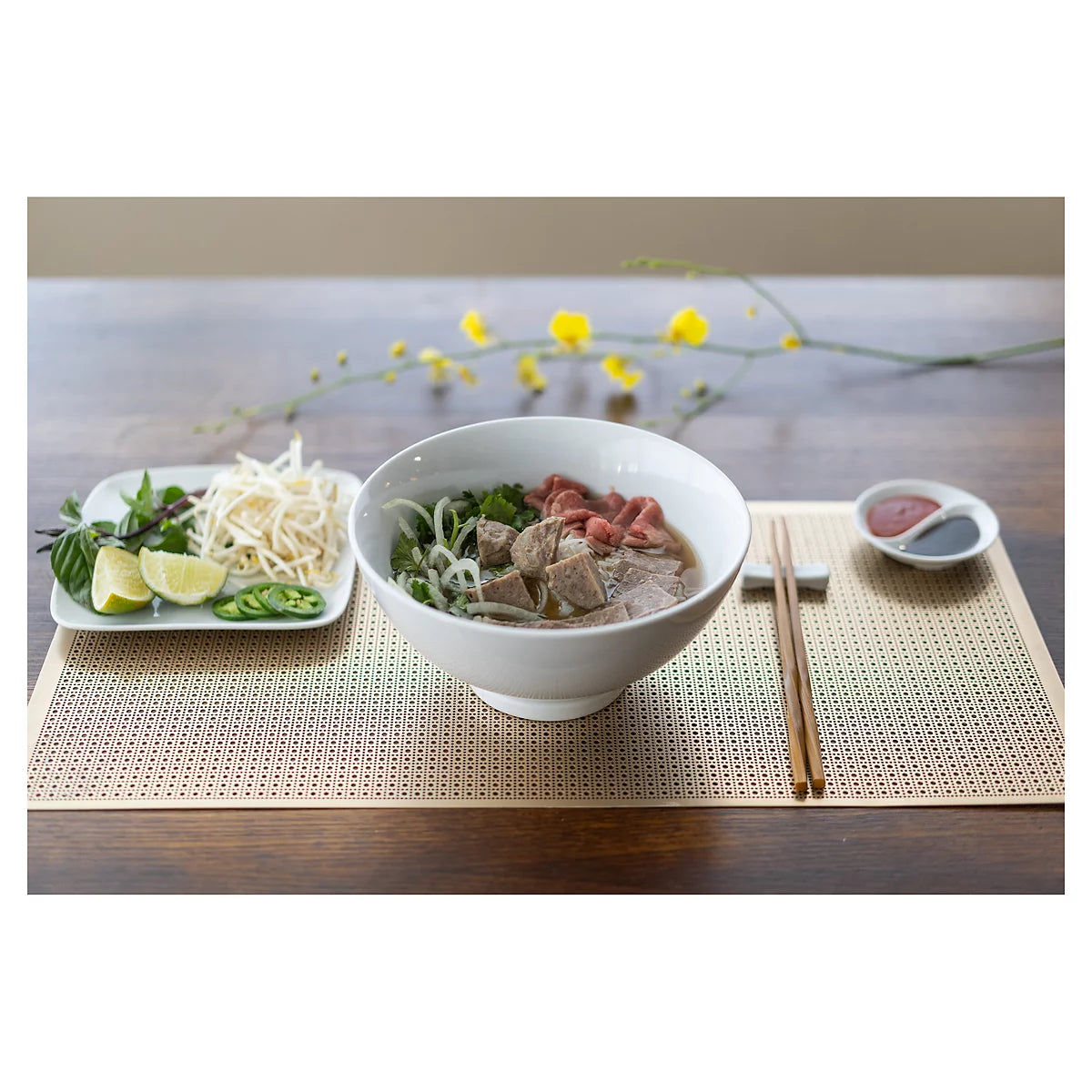 A white ceramic bowl of noodle soup with beef and greens, served on a woven beige placemat with chopsticks, a side plate with lime and bean sprouts, and a small dipping dish on a wooden table.