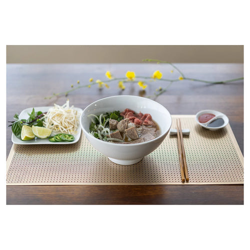 A white ceramic bowl of noodle soup with beef and greens, served on a woven beige placemat with chopsticks, a side plate with lime and bean sprouts, and a small dipping dish on a wooden table.