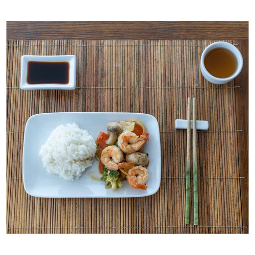 A bamboo placemat with a drink, soy dish, plate of food, and chopsticks on a chopstick rest.