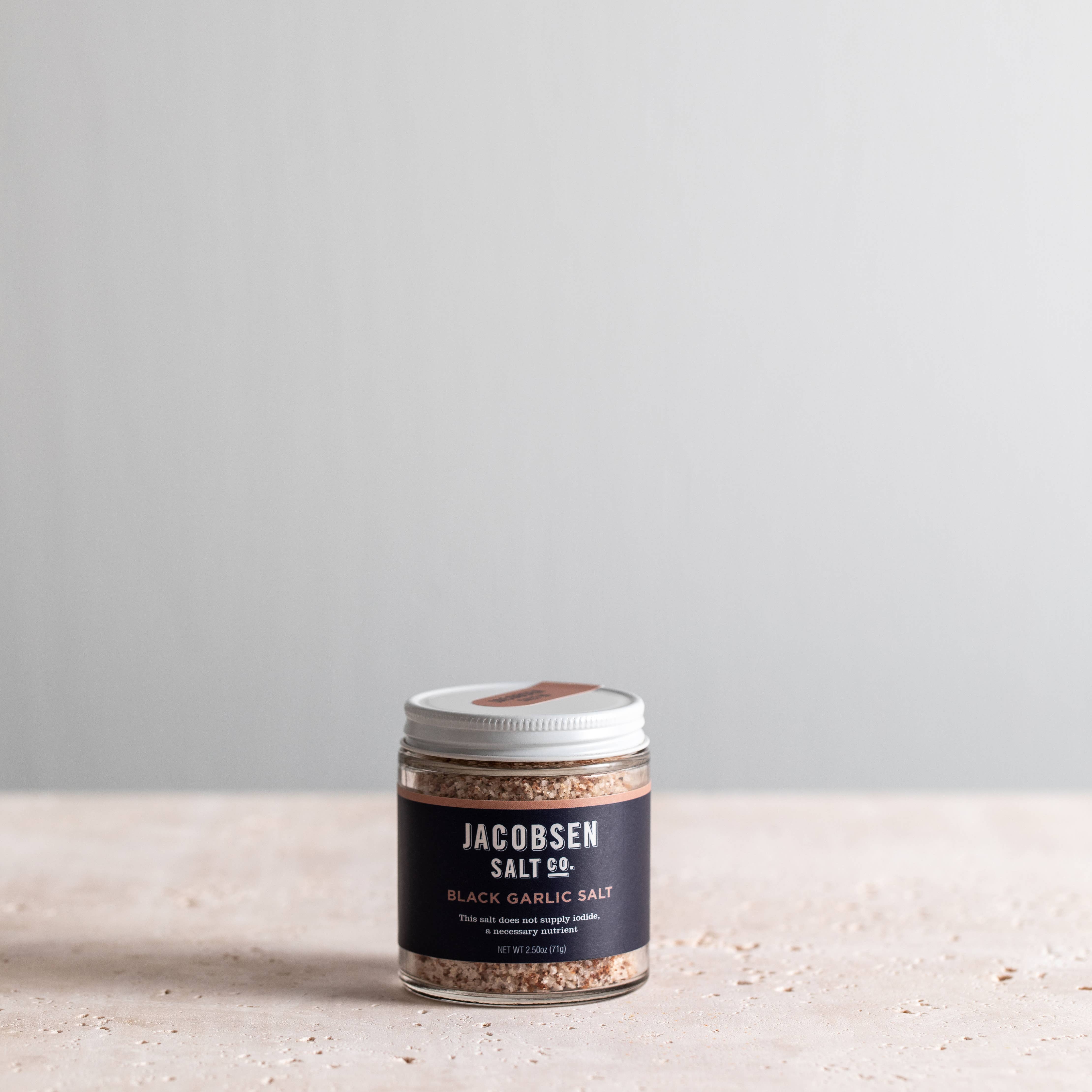 A jar of Jacobsen Salt Co. Black Garlic Salt infused sea salt, placed on a light surface.